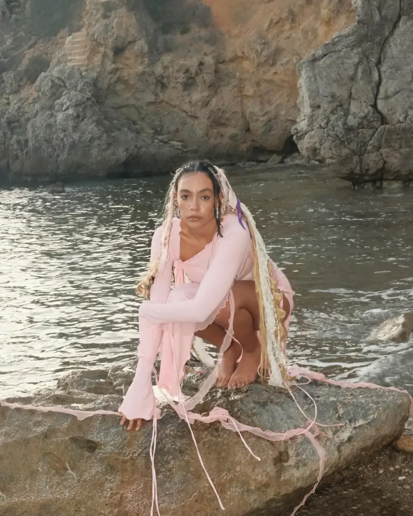Modelo con vestido rosa y trenzas decoradas, posando en una playa rocosa.