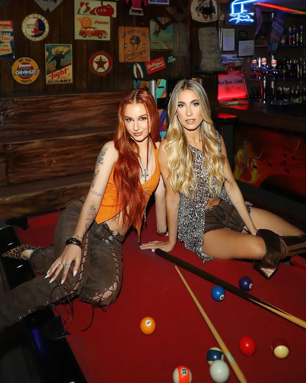Dos mujeres posando en una mesa de billar con outfits modernos y alternativos.